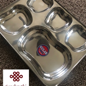 May include: A stainless steel divided serving tray with five compartments. The tray has a shiny, reflective surface and a sticker that reads "GULAB GOLD LIFE TIME". The logo for "Sanskruthi Ethnics" is in the lower left corner.