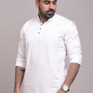 Pu&ograve; includere: Una camicia bianca a maniche lunghe con colletto alla coreana e bottoni in legno. La camicia &egrave; di colore uniforme e sembra essere realizzata in un tessuto leggero. Le maniche sono arrotolate fino ai gomiti. &Egrave; visibile anche un orologio.