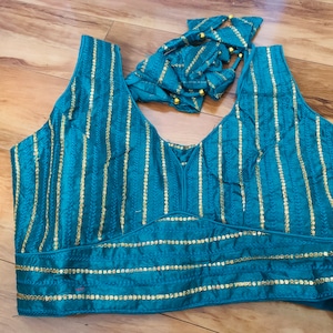 May include: Teal-colored sleeveless top with vertical gold stripes. The top features a V-neck and a decorative band at the waist. A matching teal tie with gold accents is also included. The fabric has a subtle sheen.