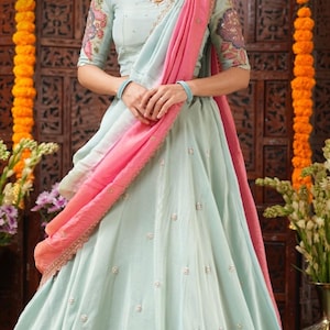May include: A light blue lehenga with delicate pearl embellishments. The outfit includes a matching top with embroidered sleeves and a pink and light blue dupatta. The text "Bullion Knot" is visible on the skirt. The code is BK1223N and the sizes are S to XXL.