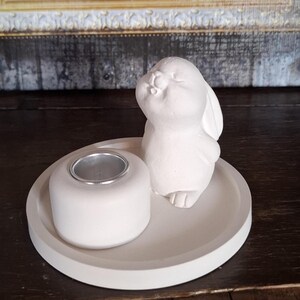 Könnte beinhalten: Weiße Keramik-Hasenfigur mit einem Teelichthalter. Der Hase sitzt auf einem runden weißen Keramiktablett.