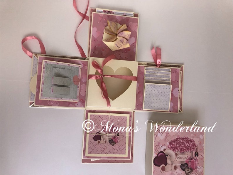 Op de afbeelding: Een handgemaakte, meerlagige explosiedoos in roze en cr&egrave;me tinten, versierd met hart- en stippenpatronen. Voorzien van een hartvormige uitsparing, uittrekbare lipjes en decoratieve elementen. Inclusief een kleine kaart met een teddybeer en bloemenontwerp.