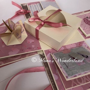 Op de afbeelding: Een handgemaakte explosiebox met een cr&egrave;mekleurige buitenkant en roze accenten. De doos heeft een hartvormige uitsparing en is vastgebonden met een roze lint. De binnenkant onthult lagen roze en bedrukt papier, met decoratieve elementen.