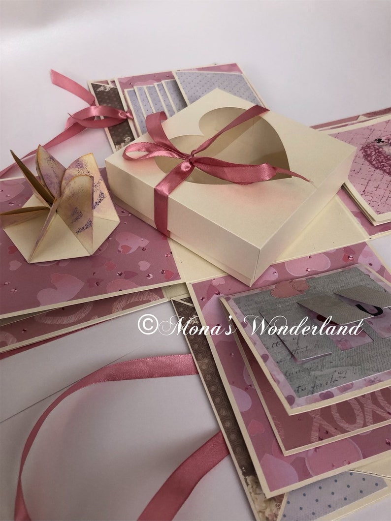 Op de afbeelding: Een handgemaakte explosiebox met een cr&egrave;mekleurige buitenkant en roze accenten. De doos heeft een hartvormige uitsparing en is vastgebonden met een roze lint. De binnenkant onthult lagen roze en bedrukt papier, met decoratieve elementen.