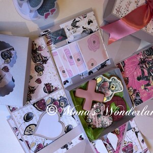 May include: An open gift box with various decorative elements. The box features patterned paper with hearts, balloons, and scooter illustrations. Inside are cookies, cards, and a pink ribbon. The box is white and pink with a heart-shaped cutout.