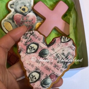 May include: A box of decorated cookies, including a teddy bear holding a heart, a pink X, and a heart-shaped cookie with a pink background and black text. The cookies are on green tissue paper.
