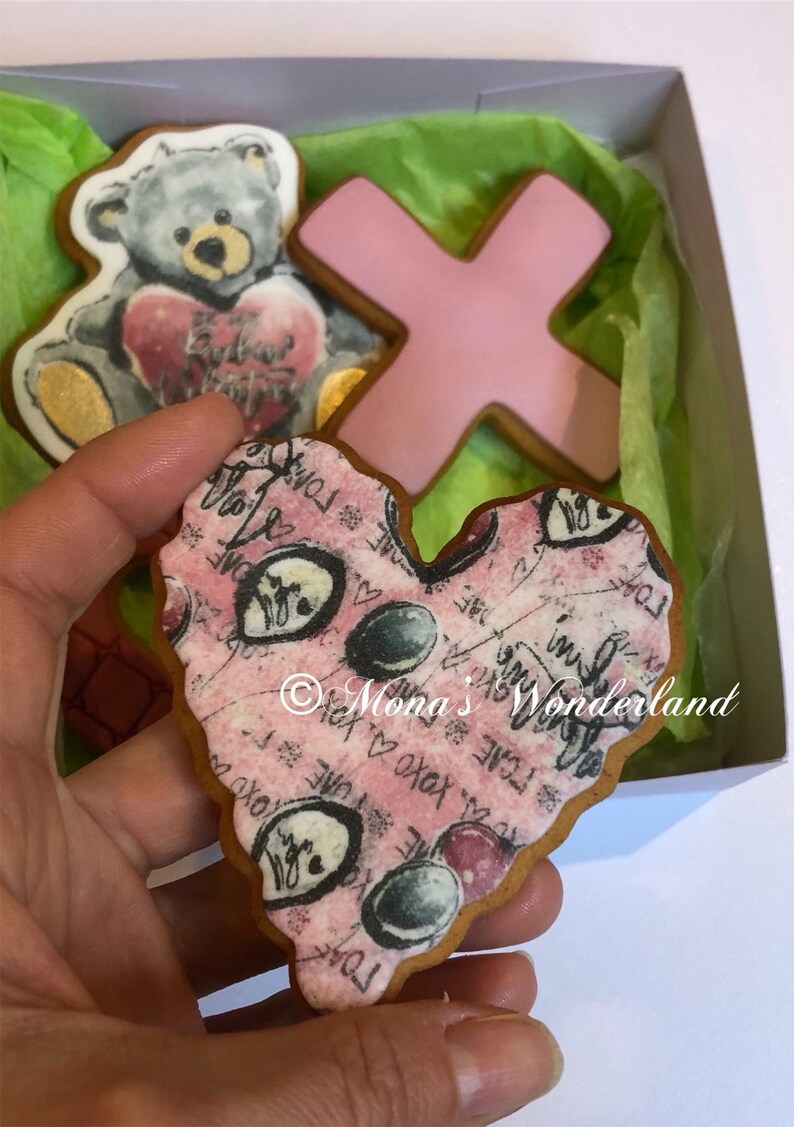 May include: A box of decorated cookies, including a teddy bear holding a heart, a pink X, and a heart-shaped cookie with a pink background and black text. The cookies are on green tissue paper.