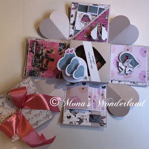 May include: A handmade explosion box with various decorative elements. The box features heart-shaped accents, patterned paper, and a large pink ribbon. The box is decorated with hearts, flowers, and other embellishments. The box is a unique gift idea.