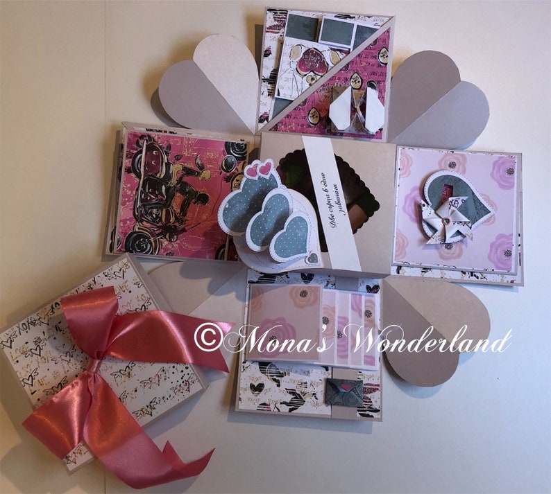 May include: A handmade explosion box with various decorative elements. The box features heart-shaped accents, patterned paper, and a large pink ribbon. The box is decorated with hearts, flowers, and other embellishments. The box is a unique gift idea.