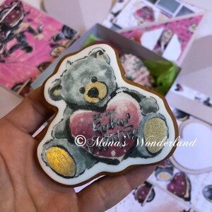 May include: A teddy bear-shaped cookie with a gray body and gold accents, holding a pink heart with the text "Be My Rockin' Valentine" and "Mona's Wonderland". The cookie is on a white background with pink boxes.