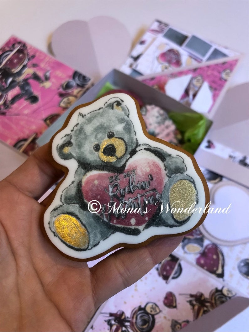 May include: A teddy bear-shaped cookie with a gray body and gold accents, holding a pink heart with the text "Be My Rockin' Valentine" and "Mona's Wonderland". The cookie is on a white background with pink boxes.