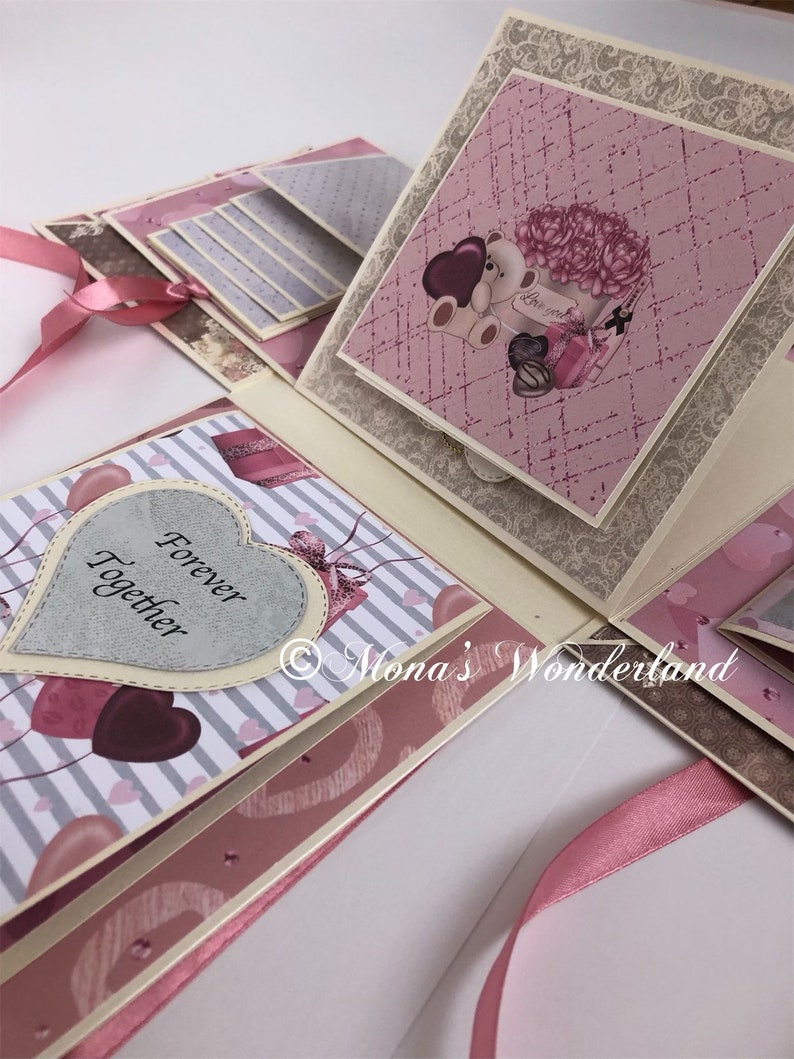 Op de afbeelding: Een open, meerlagig scrapbook met roze, grijze en cr&egrave;mekleurige papieren. Het scrapbook heeft hart- en bloemmotieven, met de tekst "Forever Together" op een hartvormig element. Een roze lint is zichtbaar.