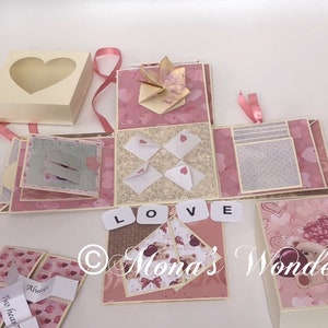 Op de afbeelding: Een handgemaakte explosiedoos met een hartvormig venster op het deksel. De doos is versierd met roze en cr&egrave;mekleurig papier, origami en kleine enveloppen met hartdetails. Het woord "LOVE" is gespeld met lettertegels. Lintjes en decoratieve elementen zijn inbegrepen.