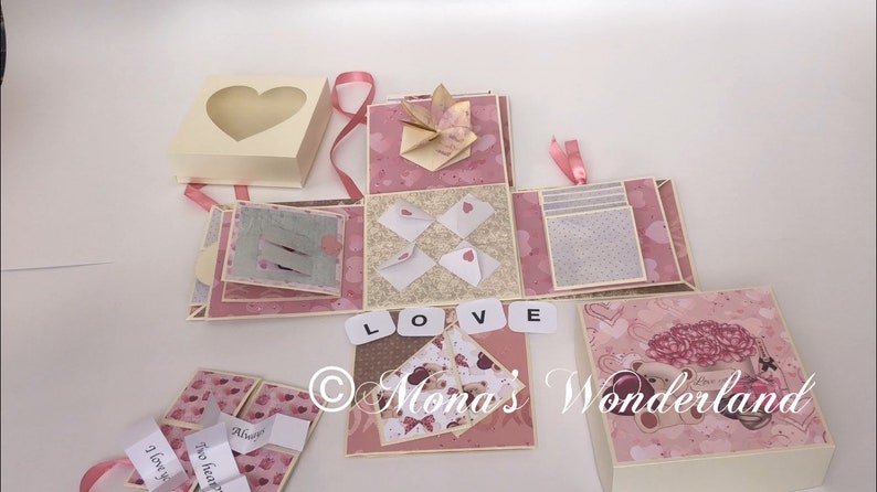 Op de afbeelding: Een handgemaakte explosiedoos met een hartvormig venster op het deksel. De doos is versierd met roze en cr&egrave;mekleurig papier, origami en kleine enveloppen met hartdetails. Het woord "LOVE" is gespeld met lettertegels. Lintjes en decoratieve elementen zijn inbegrepen.