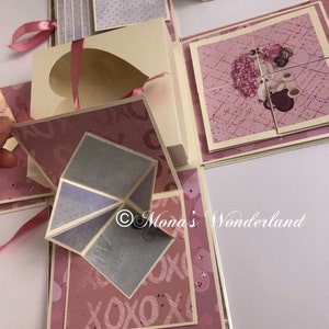 Op de afbeelding: Een handgemaakte explosiebox met meerdere lagen en compartimenten. De box heeft een hartvormige uitsparing, roze linten en diverse papieren met patronen in roze, cr&egrave;me en grijstinten. Een puzzel met een teddybeer-afbeelding is inbegrepen. De tekst "Mona's Wonderland" is zichtbaar.