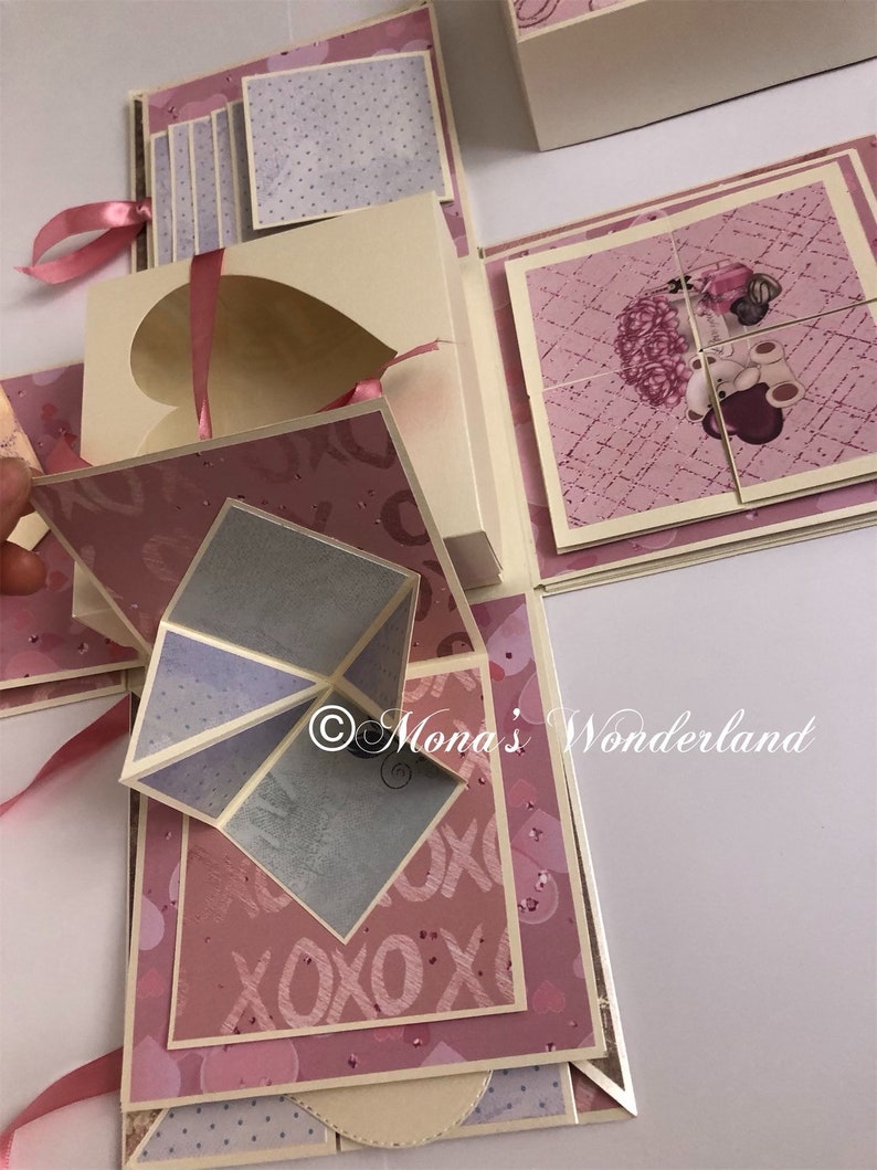 Op de afbeelding: Een handgemaakte explosiebox met meerdere lagen en compartimenten. De box heeft een hartvormige uitsparing, roze linten en diverse papieren met patronen in roze, cr&egrave;me en grijstinten. Een puzzel met een teddybeer-afbeelding is inbegrepen. De tekst "Mona's Wonderland" is zichtbaar.