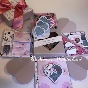 May include: A handmade scrapbook gift box with a pink bow, featuring heart-shaped elements and floral patterns. The box opens to reveal multiple layers with decorative paper and heart-shaped embellishments. Text on the box reads "Two hearts in one".