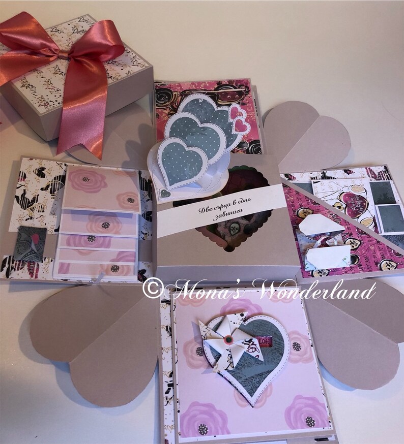 May include: A handmade scrapbook gift box with a pink bow, featuring heart-shaped elements and floral patterns. The box opens to reveal multiple layers with decorative paper and heart-shaped embellishments. Text on the box reads "Two hearts in one".