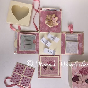 Op de afbeelding: Een handgemaakte explosiedoos met een hartvormig venster op het deksel. De doos is roze en cr&egrave;me met hartpatronen en bevat kleine kaartjes met tekst. Een teddybeer en bloemen versieren de doos.
