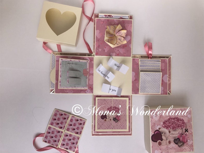 Op de afbeelding: Een handgemaakte explosiedoos met een hartvormig venster op het deksel. De doos is roze en cr&egrave;me met hartpatronen en bevat kleine kaartjes met tekst. Een teddybeer en bloemen versieren de doos.