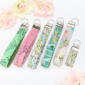 May include: Six fabric wrist keychains with silver-tone fittings. The keychains display diverse patterns, including a fairy design, floral prints, and a star pattern. The keychains are in shades of pink, green, and white.