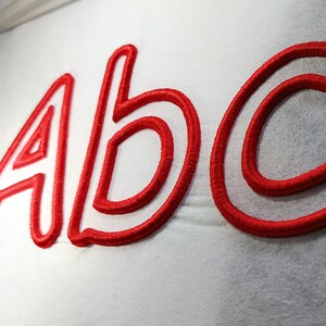 May include: Red embroidered letters "ABC" on a white fabric background.