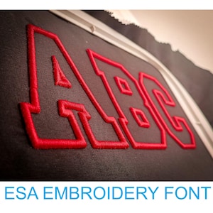 Könnte beinhalten: Nahaufnahme von roten gestickten Buchstaben "ABC" auf einem dunkelgrauen Stoff. Die Buchstaben sind weiß umrandet und haben einen leicht erhabenen Look.  ESA Embroidery Font.
