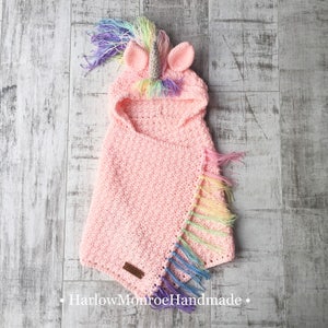 May include: Pink crocheted unicorn hooded cape with rainbow fringe and a unicorn horn on the hood.  The cape is laid flat on a wooden surface.  The text "HarlowMonroeHandmade" is visible at the bottom of the image.