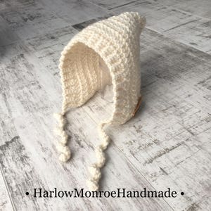 May include: A white crocheted baby bonnet with long ties. The bonnet has a pointed top and a simple design.