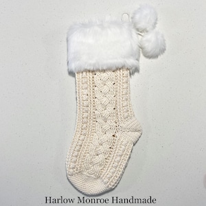Peut inclure: Une chaussette de Noël blanche en crochet avec un revers en fausse fourrure blanche et deux pompons blancs. La chaussette a un motif en tricot torsadé.