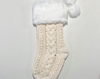 May include: A white crocheted Christmas stocking with a white faux fur cuff and two white pom-poms. The stocking has a cable knit pattern.