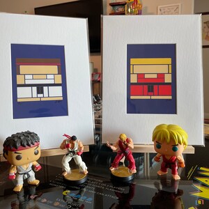Pode incluir: Duas impress&otilde;es emolduradas de retratos de personagens pixelados da s&eacute;rie de videogames Street Fighter. As impress&otilde;es s&atilde;o exibidas em uma mesa com quatro figuras dos personagens Ryu, Ken, Chun-Li e Guile. As figuras est&atilde;o todas em poses de luta.