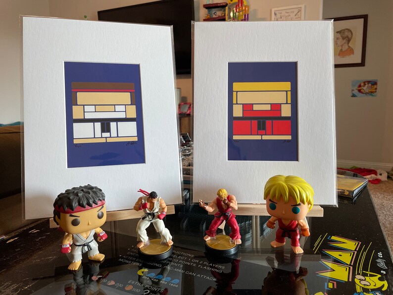 Pode incluir: Duas impress&otilde;es emolduradas de retratos de personagens pixelados da s&eacute;rie de videogames Street Fighter. As impress&otilde;es s&atilde;o exibidas em uma mesa com quatro figuras dos personagens Ryu, Ken, Chun-Li e Guile. As figuras est&atilde;o todas em poses de luta.