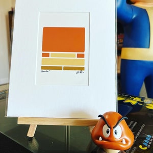 May include: A framed print of a minimalist geometric design in shades of orange, yellow, and brown. The print is titled "Goomba" and is signed by the artist. The print is displayed on a small wooden easel.