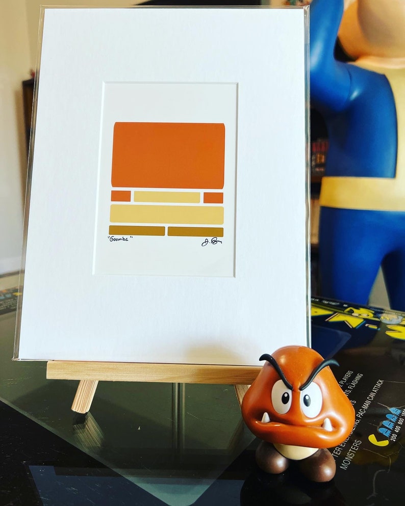 May include: A framed print of a minimalist geometric design in shades of orange, yellow, and brown. The print is titled "Goomba" and is signed by the artist. The print is displayed on a small wooden easel.