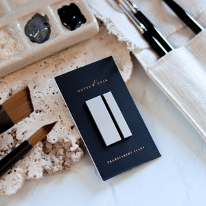May include: Two gray rectangular sticky notes on a black card with the text "NOTES OF DUSK" and "TRANSPARENT FLAGS". The card is on a light brown textured surface with two paint brushes and a white ceramic paint palette.