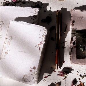 May include: Two white notebooks with floral designs and the word "NOTES" printed on the front. A gold pen and a pink notebook with a green ribbon are also visible on a white surface with dried flower petals.