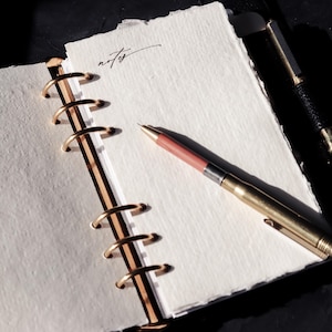 May include: A white notebook with a gold spiral binding and the word "notes" written on the first page. A gold pen with a pink eraser is lying on the open notebook.