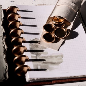 May include: A white notebook with a grid pattern and a watercolour design. The notebook is open and has a gold binder with rings. A small pouch with gold rings is on top of the notebook.