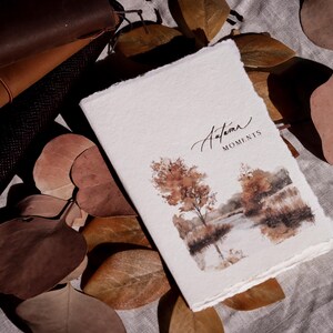 May include: A white journal with the words "Autumn Moments" in script, featuring a watercolour landscape of trees and a lake. Brown leaves and a leather notebook are in the background.