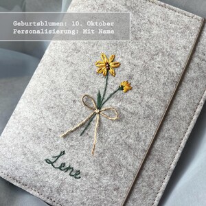 Könnte beinhalten: Ein hellgrauer Filz-Notizbuchumschlag mit einer gestickten gelben Blume, einer Schleife aus Bindfaden und dem Namen Lene in dunkelgrünem Garn. Der Text Geburtsblumen: 10. Oktober Personalisierung: Mit Name ist auf dem Bild zu sehen.