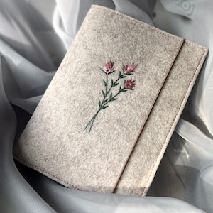 Könnte beinhalten: Ein Notizbuchumschlag aus hellbeigem Filz mit einer zarten, handgestickten Verzierung aus drei kleinen, rosafarbenen, kleeartigen Blüten mit grünen Stielen und Blättern. Der Umschlag wird durch ein schmales, vertikales Gummiband verschlossen.