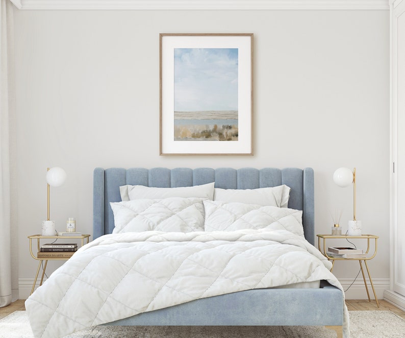May include: A light blue upholstered bed with a white quilted duvet and two white pillows. The bed is in a bedroom with a framed landscape photograph on the wall and two gold side tables with lamps.