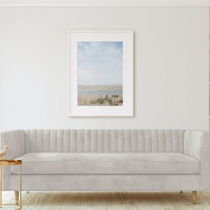 May include: A light gray tufted sofa with a gold base sits in a living room with a framed landscape photograph on the wall and a white bookshelf with decorative objects on the right side.
