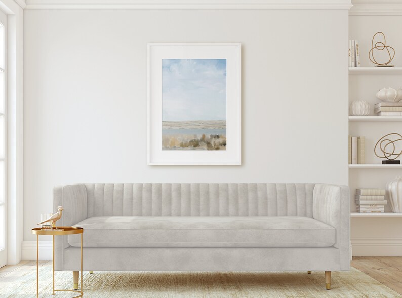 May include: A light gray tufted sofa with a gold base sits in a living room with a framed landscape photograph on the wall and a white bookshelf with decorative objects on the right side.