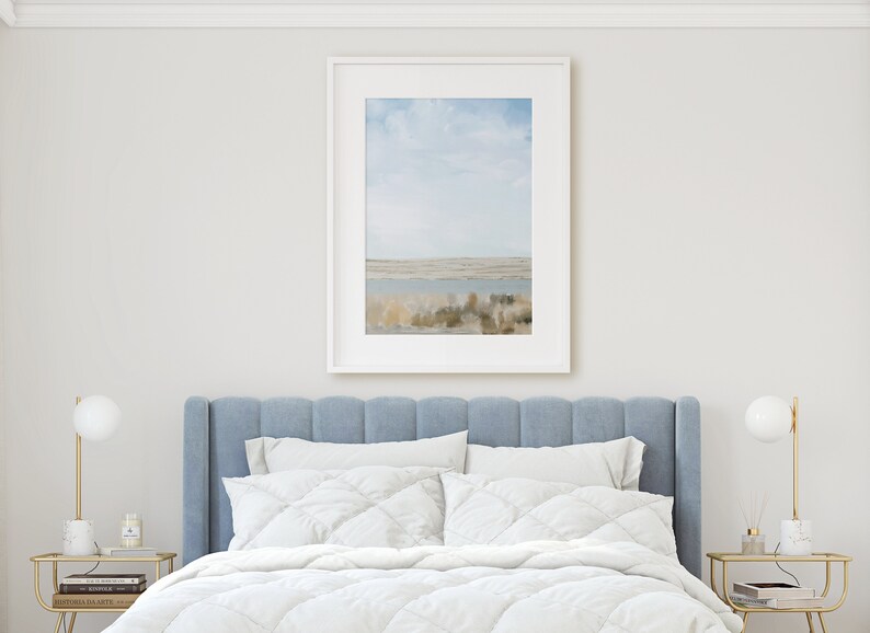May include: A white bed with a blue headboard and white bedding. A framed print of a landscape with a body of water and a cloudy sky hangs above the bed. Two gold lamps with white shades are on nightstands on either side of the bed.