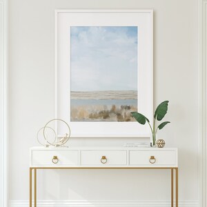 May include: White console table with three drawers and gold metal legs. The table has a framed painting of a landscape with a blue sky and water. A potted plant and decorative objects are on top of the table.