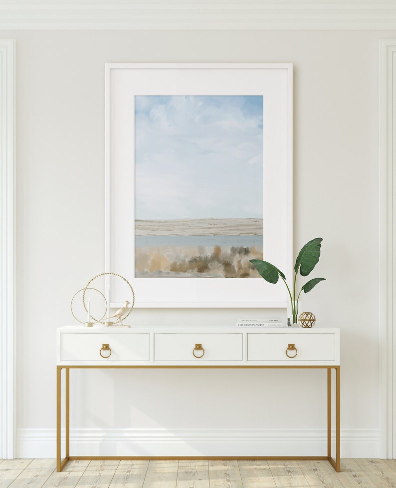 May include: White console table with three drawers and gold metal legs. The table has a framed painting of a landscape with a blue sky and water. A potted plant and decorative objects are on top of the table.