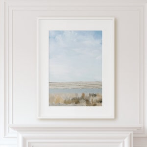 May include: A framed print of a landscape painting with a blue sky, white clouds, and a body of water in the distance. The foreground features a field of brown and green grasses.