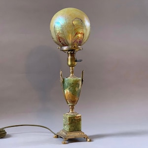 May include: A green and gold table lamp with a round iridescent glass shade. The lamp has a base made of green marble with gold accents.